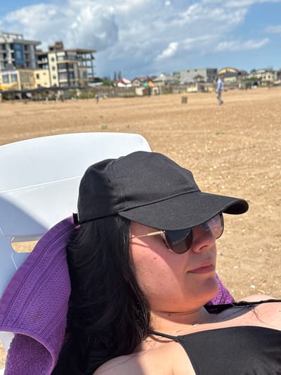 The image shows a woman lying on a white beach chair on a sandy beach, with a purple towel draped over the chair behind her head. She is wearing a black baseball cap, sunglasses, and a black bikini top. The background depicts buildings and the sky with clouds. The scene is consistent with a beach resort area in Derbent, Russia, likely during daytime with fair weather. The woman appears to be relaxed and enjoying the sun.