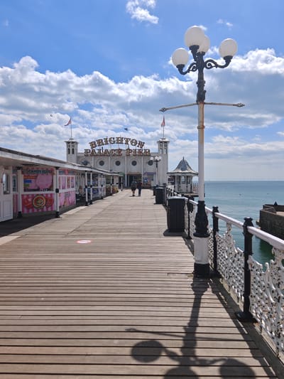 The image depicts Brighton Palace Pier in Brighton, United Kingdom. The pier is made of wooden planks and has shops selling candy floss on the left side. In the background, there is the Brighton Palace Pier building with flags on top. A tall, ornate lamppost stands on the right. The sky is blue with fluffy white clouds. The sea is visible to the right of the pier, with a wind farm visible on the horizon. Two people are walking further down the pier. The image shows an empty pier with an absence of people.