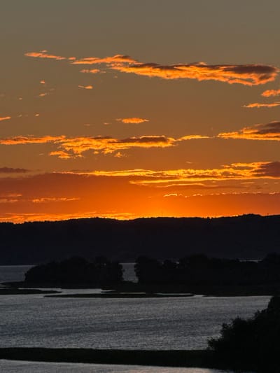 The image depicts a sunset over a body of water, likely a river or bay, with a silhouetted treeline forming the horizon. The sky is filled with vibrant orange and yellow hues, with scattered clouds catching the last rays of sunlight. In the foreground, the water reflects the colors of the sky and appears calm. The overall scene is tranquil and evokes the end of the day. The provided crops highlight the detail of the clouds and the gradient of colors in the sky. Without further information, the specific location within the city of Bolgar, Russia, cannot be determined, but the landscape suggests a natural setting within or near the city.