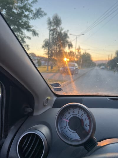 The image is taken from inside a car, looking out through the windshield. The dashboard is visible in the foreground, with a tachometer showing approximately 1000 rpm. The windshield is dirty and streaked, with several scratches visible. Outside the car, the sun is setting, casting a warm, orange glow across the scene. The sun's rays are reflecting off the windshield, creating a lens flare effect. A road is visible, with a few cars driving on it. Trees and houses line the side of the road. The sky is a clear blue with some haze. The location appears to be a suburban or rural area. The overall impression is that of a peaceful evening drive. The image could be of the area near Delchevo, North Macedonia, as suggested.