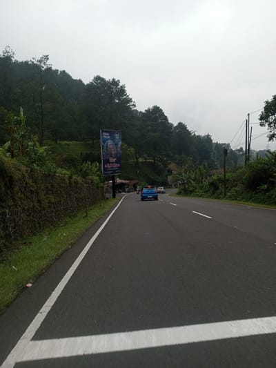 The image shows a road in a mountainous area, possibly in Mae Chan District, Thailand, although without further information, it is impossible to say with certainty. The road is paved and has white lines marking the lanes. A blue car is driving on the road, with another car further ahead. There is a large billboard on the left side of the road, advertising "Glass House Roof Park" along the side of a stone wall, and behind it are trees and greenery. The sky is overcast and gray. Power lines are visible on the right side of the road, surrounded by dense vegetation.
