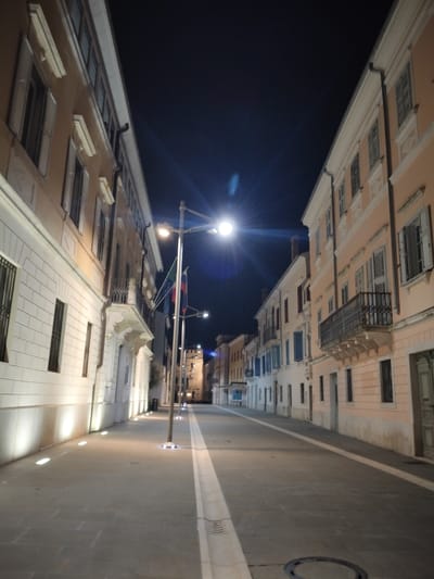 A deserted street at night is lined with buildings of different styles. Streetlights illuminate the scene, casting shadows and reflections on the cobblestone pavement. A white stripe runs down the center of the road. Flags are visible near the streetlights. The buildings feature windows with closed shutters. A balcony adorns one of the buildings on the right. The sky above is dark.
