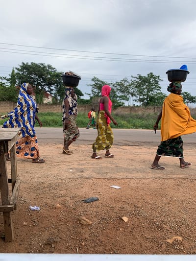 In Gbeleko, Nigeria, four women are walking single file along a road. The woman at the front is wearing a yellow shawl, a green and black skirt, and sandals, and she has a black bucket on her head. The second woman is wearing a pink top, a yellow and green patterned skirt, and sandals, and she is carrying something green and red in her hands. The third woman has a black bucket on her head, and she is wearing a blue and white patterned top, a tan skirt, and sandals. The last woman is wearing a blue and brown patterned dress and sandals. The road is unpaved and there is a wooden structure in the foreground on the right side of the image. There are trees and bushes in the background, and power lines overhead.