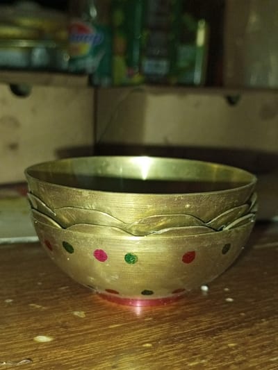A stack of three brass-colored metal bowls rests on a worn wooden surface in Longmiang, Laos. The bottommost bowl, partially visible, is adorned with rows of alternating red and green circular dots. The upper two bowls, nested inside each other, display scalloped edges around their rims. All bowls exhibit a dull, somewhat tarnished metallic sheen with visible surface abrasions. The wooden surface below features discernible grain patterns and scattered light-colored particulate matter. The background is out of focus, revealing blurred objects on shelves, including indistinct containers, one primarily green and another with blue and white elements. No individuals, actions, or interactions are depicted.