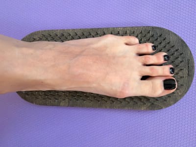 A person's left foot, with black nail polish on the toes, rests on a wooden board with protruding nails. The board is placed on a purple, textured mat. The scene appears to be indoors, likely in a residential setting. There are no discernible landmarks or contextual clues that would indicate a specific location in Novoaleksandrovka, Russia.