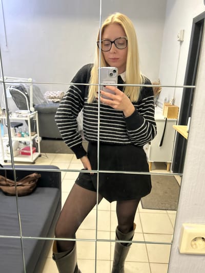 The image depicts a blonde female taking a mirror selfie indoors in Voskresensk, Russia. The subject, visible from head to upper shin, wears dark-framed eyeglasses, a black and white horizontally striped sweater over a white collar, a black skirt, black sheer tights, and dark knee-high boots. She holds a light-colored smartphone with her left hand, displaying dark nail polish, while her right hand is partially obscured, resting in her skirt pocket. The mirrored reflection reveals a room with light gray walls and light-colored tiled flooring. To the subject's left, a dark-colored sofa with a brown bag rests adjacent to a white multi-tiered rolling utility cart containing various small items. To the subject's right, a white cabinet with a wooden top and decorative elements is visible, along with a dark door frame and a light-colored electrical outlet mounted on the wall.
