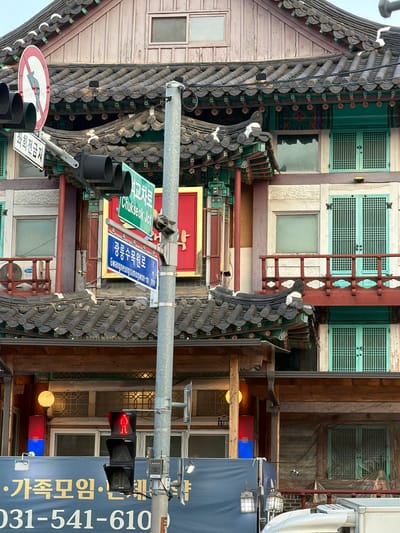 A traffic light with a red walking figure is visible on a pole in front of a traditional Korean building. The building features intricate tile work on its roof and decorative paintings on its eaves. Green-framed windows with lattice designs are visible on the building's facade. A sign with Korean text and a street name "Gwangneungsumogwon-ro" is mounted on the pole. Below the traffic light, a blue banner with Korean text and a phone number "031-541-610" is displayed. The scene is an outdoor urban environment.