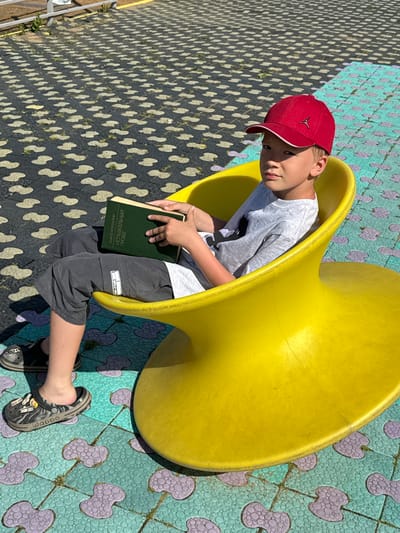 A child sits inside a yellow playground structure, reading a book. The child wears a red baseball cap, a gray t-shirt, gray shorts, and Crocs-style shoes. The book has Cyrillic text on its cover. The playground surface is patterned with interconnected designs in shades of teal, purple, and gray. Another surface with a different pattern, black with light shapes, lies nearby. The scene is in Синьково, Russia.