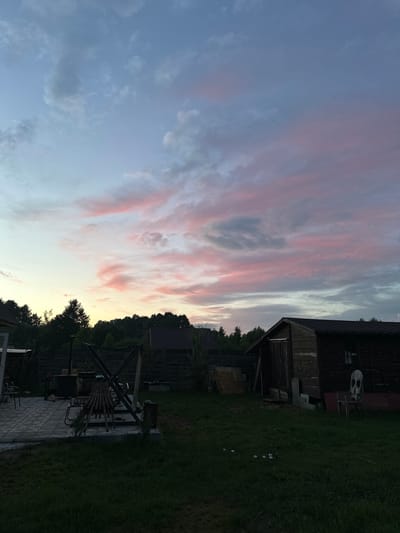 The image depicts a twilight scene in a rural backyard, likely in the Тумілавіцкі сельскі Савет, Belarus. The sky is filled with dramatic clouds tinged with pink and purple hues, suggesting the end of a day. In the foreground, a grassy lawn stretches towards a paved patio area. On the patio, there are several chairs, a barbecue grill, and a wooden swing set. To the right of the patio, a rustic wooden shed stands, its dark wood contrasting with the lighter tones of the sky. A chair with a skull design is visible near the shed. Several small, round, white objects, possibly balls or decorative elements, are scattered on the grass. The background is a line of dark trees, silhouetted against the colorful sky, with the roofs of houses peeking through them. The overall impression is one of a peaceful, domestic setting bathed in the soft light of sunset. There are no people visible in the image.