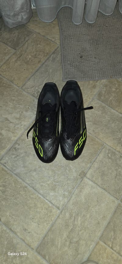 The image displays two black athletic shoes, likely soccer cleats, positioned on a light brown tiled floor. The shoes are oriented lengthwise, with one shoe pointing towards the top of the frame and the other pointing towards the bottom, creating a symmetrical arrangement. Both shoes feature prominent bright yellow-green "F50" lettering on their sides and matching stripe patterns.
To the left of the shoes, a grey, textured, woven floor mat is visible. Further to the far left, a light grey, possibly upholstered, object is partially seen, suggesting furniture or another soft furnishing. The tiled floor has visible grout lines and exhibits a subtle mottled texture.
No activity is taking place; the shoes are stationary. The scene appears to be an indoor setting, likely a home, based on the flooring and mat. The lighting is uniform, not indicating a specific time of day. The shoes appear to be in good condition.
Visible text includes "F50" on the side of both shoes. A "Galaxy S24" watermark is present in the bottom right corner of the image, indicating the device used for capture. The provided location context is Laval, Canada, though no specific visual cues in the image confirm this.
