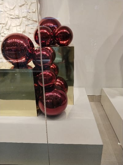 A stack of reflective, maroon-colored spheres sits on a glossy, gold-toned cubic structure. The arrangement rests on a white platform. A textured, white material is visible in the background. The scene appears to be behind glass, creating a mirrored effect on the orbs and geometric shapes. To the right, another white structure and the floor are visible.