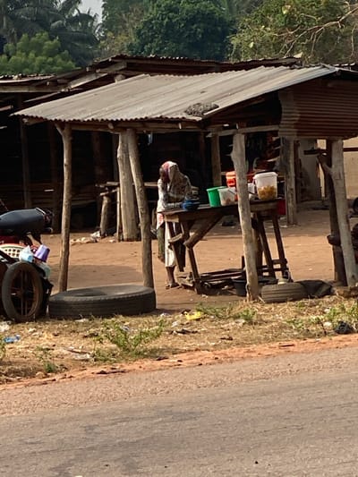 This image depicts a roadside scene in Okura, Nigeria. The foreground shows a paved road surface adjacent to an unpaved, reddish-brown dirt area. This dirt area contains sparse dry vegetation and scattered refuse, including plastic bags and debris. A single black vehicle tire lies flat on the ground in the mid-left.
At the center, a makeshift stall or shop structure is visible, featuring a weathered corrugated metal roof supported by multiple rough-hewn wooden poles. Under this structure, one individual, possibly a woman, stands or leans behind a wooden table. Her head is covered with a patterned fabric, and she wears a light-colored, long garment. Her face is obscured. On the wooden table in front of her are several containers: a blue bowl, a green bucket, a white bucket, a reddish-orange container, and a translucent container holding numerous light-colored spherical items, likely food.
To the left of the stall, a portion of a dark-seated motorbike with a spoked wheel is visible, appearing to be parked. Behind the primary stall, other similar rustic structures with corrugated roofs and wooden supports are partially discernible. The background features dense green foliage and trees under what appears to be natural daylight.