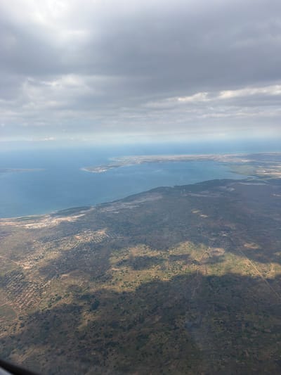 An aerial view captures a coastal landscape in Metuge, Mozambique. The image is dominated by a vast expanse of blue ocean on the left, meeting a sandy coastline. Beyond the coast, the land transitions into a varied terrain with areas of vegetation, scattered settlements, and visible roads. In the upper portion of the image, a cloudy sky with diffused sunlight covers the scene. The overall impression is of a developing coastal region with a mix of natural and man-made elements under a vast sky.