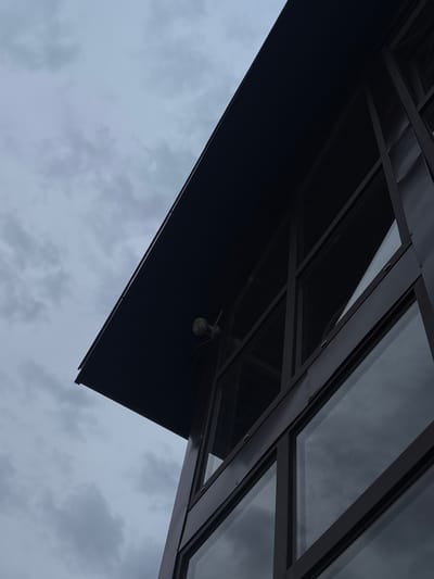 The image shows a modern building with a dark, angular roof and large windows. A security camera is visible mounted on the underside of the roof overhang. The sky is overcast with gray clouds. The architecture suggests an urban or commercial setting. The specific location in Gubakha, Russia is not discernible from the visual information provided.