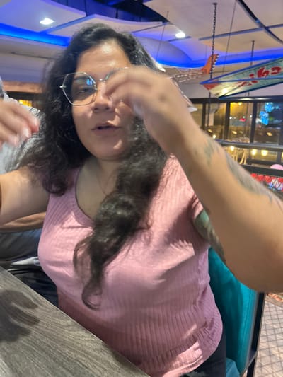 A woman with dark, curly hair and glasses is wearing a light pink shirt. She is seated at a table in what appears to be an outdoor dining area. Blue lighting accents the ceiling, and a building with numerous windows is visible in the background. The woman's arm, adorned with tattoos, is raised in the foreground, partially obscuring the view. The photo is taken at an unusual angle, making it appear tilted.