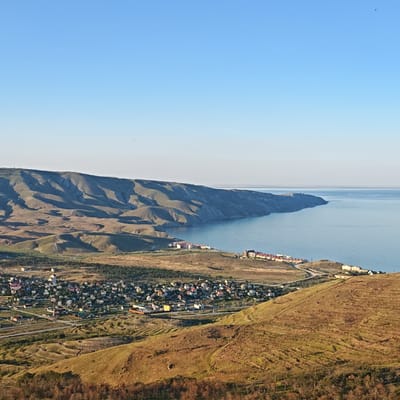 This is a high-angle, wide shot photograph taken from a hill overlooking a coastal town in Ordzhonikidzevskoye, Ukraine. The sky is a clear, pale blue with a gradient from lighter at the horizon to a deeper blue at the top. The ocean, a calm, light blue, stretches out to the horizon, meeting the sky.

In the middle ground, a town with many small houses, predominantly with red roofs, is nestled between the hills and the sea. Roads weave through the town, and a few larger buildings, possibly hotels or apartment complexes with white and red facades, are visible along the coastline. The hills surrounding the town are arid and dry, with terraced slopes in some areas, giving them a layered appearance. The colors of the hills range from dry yellow-brown grasses to muted green patches.

In the foreground, the hill continues to slope downwards, covered in dry, golden-brown vegetation and scattered trees. The light suggests it is late afternoon or early morning, casting long shadows across the landscape. There are no visible people in the frame. The scene depicts a rural coastal settlement with natural, mountainous terrain.