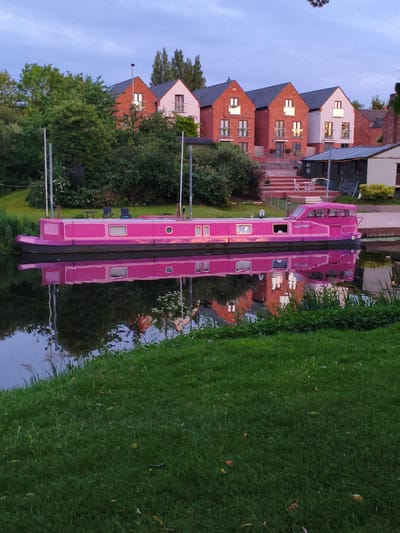 This is a horizontal, eye-level, full shot of a bright pink canal boat moored on a canal. The boat is the primary subject, positioned in the foreground, with its reflection clearly visible in the calm water. The boat is long and narrow, with several windows and a covered front section. There are two tall poles on the deck of the boat, likely for mooring.
Behind the canal boat, on the bank, is a patch of vibrant green grass. Further back, there are several modern houses with red brick and white facades, featuring prominent windows and balconies. Some of these houses appear to be illuminated from within. To the left of the houses, there is a dense line of trees and foliage. A small, dark-colored shed or outbuilding with a pitched roof and string lights is situated in front of the houses.
The sky above is a soft, dusky blue with a few wispy clouds. The overall lighting suggests it is either early morning or late evening. The scene evokes a sense of tranquility and suburban living. The canal boat's bright color stands out against the more muted tones of the background. Based on the distinctive architecture of the houses and the presence of a canal, this scene is likely located in a residential area along a waterway in Wychavon, United Kingdom, possibly a newer development that has incorporated canal-side living. There are no people visible in the image.