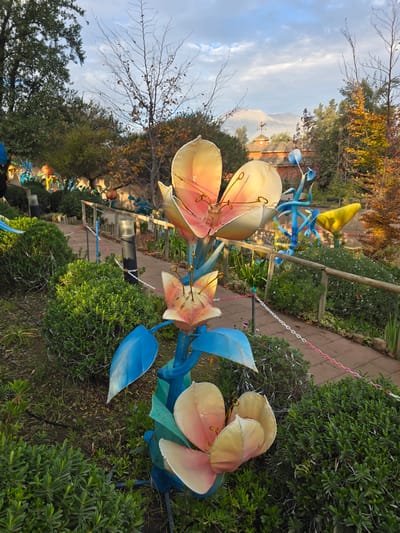 The image shows a display of large, artificial flowers in a garden setting. The flowers have pale, petal-like structures with pinkish centers and are supported by blue and green metal stems and leaves.
The garden features neatly trimmed bushes, a wooden fence along a brick path, and a variety of trees in the background. A mountain is visible in the distance.
While the image likely contains details that could lead to the conclusion that the picture was taken in Puente Alto, Chile, such as the mountain visible in the background resembling the Andes mountains visible in the region, the information needed to confirm the location is insufficient.