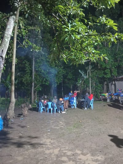 This image depicts a nighttime outdoor gathering in Butali Chegulo ward, Kenya. The scene features approximately ten to twelve individuals seated on blue plastic chairs around various low tables or surfaces on a dirt ground. Smoke or vapor is visibly rising from the left-center of the frame. The area is illuminated by artificial light sources, casting shadows, most notably a human-shaped shadow on the lower right foreground.

The environment is characterized by numerous tall trees with dense green foliage, suggesting a natural or semi-forested setting. On the far right, a simple building with a dark grey wall and a sloped roof is visible, in front of which at least three motorcycles/mopeds are parked. Tree stumps or cut wood are present on the ground near the center-left. Individuals are dressed in casual attire, some wearing hats, and appear to be engaged in conversation or social interaction. The overall ambiance is that of a communal nighttime assembly.