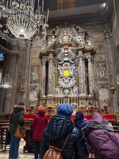 This image depicts the ornate Baroque interior of the Oratory of the Immaculate Conception, located within the Basilica di San Francesco in Ferrara, Italy. The central focus is a grand altar constructed from veined marble columns and gilded architectural elements, featuring a richly decorated central niche containing a venerated, glowing yellow icon or reliquary.
The altar is adorned with multiple candelabra holding unlit candles, smaller votive candles, and red floral arrangements, likely poinsettias, on a red altar cloth. Above the central niche, white marble sculptures of cherubs and two seated figures flank a cartouche. To the left of the altar, a wall painting depicts the Madonna and Child, with partially legible text below. To the right, another painting portrays a seated saint, explicitly identified by the inscription "S. ALPHONSUS DE LIG. EP. DOCT." (Saint Alphonsus Liguori, Bishop and Doctor), with an illegible dedication plaque below.
The high ceilings are dimly lit, illuminated primarily by two large, multi-tiered crystal chandeliers with unlit candles, one prominently hanging in the upper left foreground. Walls are decorated with pilasters and architectural details. Hanging brass lamps with red glass elements are positioned on either side of the altar.
In the foreground, five individuals are observed from behind, standing and facing the altar. All are dressed in winter attire, including puffer coats and knit hats. From left to right: one wears a dark green coat with a fur-lined hood and a purple hat; another wears a red puffer coat and an orange hat; a third wears a dark blue puffer coat and a blue hat; a fourth wears a purple puffer coat and a purple fur hat; and the fifth wears a maroon/purple puffer coat with a fur-lined hood. Two individuals carry shoulder bags: one brown bag on the far left, and another brown bag on the central figure. Wooden pews are partially visible in the lower left.