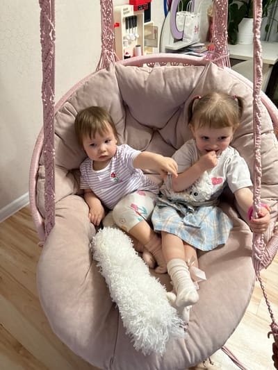 Two young children are lying together in a hanging chair. The chair is round, cushioned, and light pink, suspended by a woven pink cord. The girl on top has blonde hair in pigtails, wearing a white t-shirt, a blue plaid skirt, and white socks. She has a stuffed animal with her. The child below her has short brown hair and is wearing a striped shirt and patterned shorts. There's a white fluffy pillow beside them. The location is indoors, the environment being a domestic residence. There is a child's play kitchen set visible in the background. The image doesn't suggest any location in the city of Krapivka, Russia.
