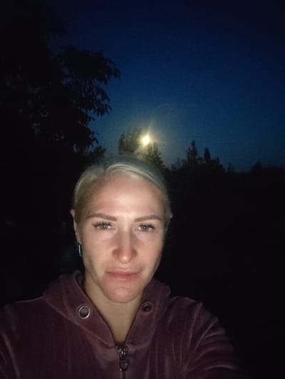 A frontal, medium close-up selfie shows a fair-skinned woman with blonde hair against a dark blue night sky with a bright, blurry light source in the upper center. The woman's face is illuminated, with light makeup including mascara and defined eyebrows. She is wearing a dark purple hooded jacket with a zipper and two silver grommets on the collar. The background is dark, with silhouettes of trees and foliage on the left and right. The location is Staiky, Ukraine.