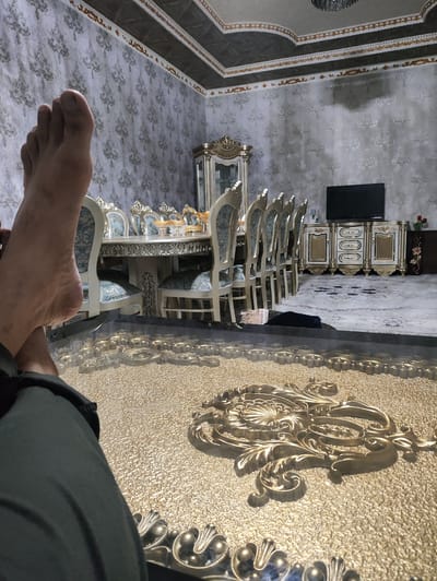 This media file depicts a luxurious interior setting, likely a dining room or living area, in Uzbekistan, possibly during the Navruz celebrations. The foreground is dominated by a close-up view of a person's bare foot and lower leg, resting on a richly decorated, golden surface that appears to be a large rug or a decorative mat with ornate, raised metallic patterns. The foot suggests a relaxed posture, implying a moment of rest or casual observation.
In the midground and background, a formal dining set is visible. This includes a long, ornate dining table with a light-colored, possibly marble or polished wood, surface. Several chairs with elegant, upholstered seating in a blue and white pattern surround the table, some featuring gold trim. To the right of the dining area, a large, ornate sideboard or cabinet is present, adorned with gold accents and decorative carvings. Above it, a flat-screen television is mounted on the wall. The walls are covered in textured wallpaper with a repeating damask or floral pattern in muted tones. The ceiling is also elaborately decorated with patterned panels and decorative borders in gold and dark tones.
The overall impression is one of opulence and comfort, with rich materials and intricate detailing throughout the space. The lighting appears to be artificial, suggesting it could be evening or the interior is well-lit regardless of the time of day. No specific event is explicitly shown, but the formal setting and the presence of Navruz decorations in the broader context could indicate a celebratory gathering or a well-appointed home. No visible text is present in the image.