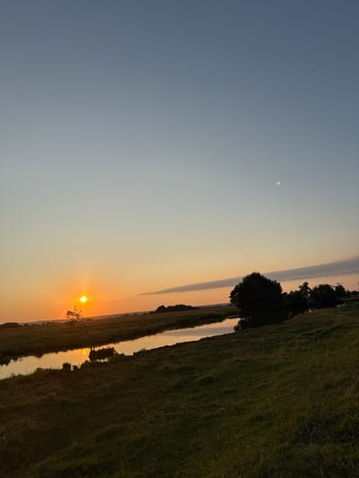 The image depicts a rural landscape at sunset in Boriskino, Russia. The sky transitions from a pale blue at the top to warm orange hues near the horizon. The sun, a bright orange orb, is setting on the left side of the frame, casting a golden reflection on a calm river that winds through a grassy field. The riverbanks are lined with vegetation and a few scattered trees. On the right side of the river, a cluster of darker trees stands in silhouette against the fading light. In the far distance, a faint outline of hills or a treeline is visible. There are no people or animals visible in the scene. The overall impression is one of tranquility and natural beauty as day transitions into night.