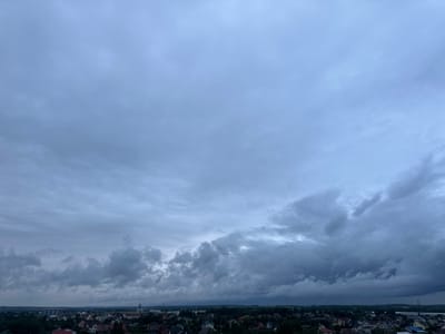 The image captures an aerial view of a suburban area, likely in Шчомысліцкі сельскі Савет, Belarus, under a heavily overcast sky. The majority of the frame is filled with dense, grey clouds, with some areas appearing darker and more ominous, suggesting impending rain or a storm. Below the sky, a dense residential area is visible, characterized by a multitude of houses with varying roof colors, predominantly red and brown. The houses are clustered together, interspersed with numerous trees that form a green canopy over the neighborhood. In the distance, industrial structures, including what appear to be silos and a tower with visible infrastructure, rise above the treeline, hinting at the proximity of more developed or industrial zones. The overall atmosphere is somber and muted due to the overcast conditions, with no people or specific actions clearly discernible in the scene.