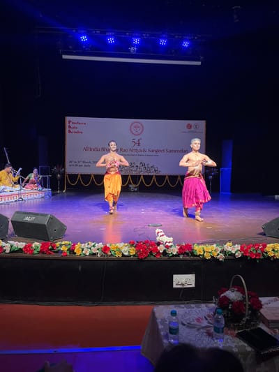 Two men in traditional Indian clothing are performing a dance on a stage. They are both wearing bright colored costumes and are holding their arms in the air. The stage is decorated with flowers and the banner behind them says "54th All India Bhar Rao Nritya & Sangeet Sammelan" and "Celebrating the Soul of Indian Classical Arts". The stage is lit with bright lights and the audience is seated in the dark. The location is the Pracheen Kala Kendra, Sector 35, Chandigarh, India, with the coordinates (latitude: 30.733, longitude: 76.789).  There is a microphone and a speaker on the stage to the left of the performers, and a basket of flowers is on the table in the foreground.