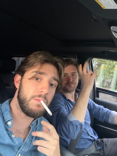 Two men are in a car, likely a luxury SUV, with the roof interior visible and the exterior showing trees and streetlights through the window. The man in the foreground, positioned on the left side of the frame, is looking at the camera and holding a cigarette in his mouth. He has brown hair, a beard, and is wearing a blue denim shirt. The man in the background, on the right, is looking towards the camera and has his right hand raised, making a gesture with two fingers extended. He has brown hair and is wearing a blue patterned shirt. Both men appear to be of Caucasian descent. The interior of the car is dark, with the visible part of the dashboard indicating a modern vehicle. The lighting suggests it is daytime, possibly late afternoon given the ambient light. The location cannot be determined from the image alone, but the mention of Cuautitlán Izcalli, Mexico, suggests the context of the image.