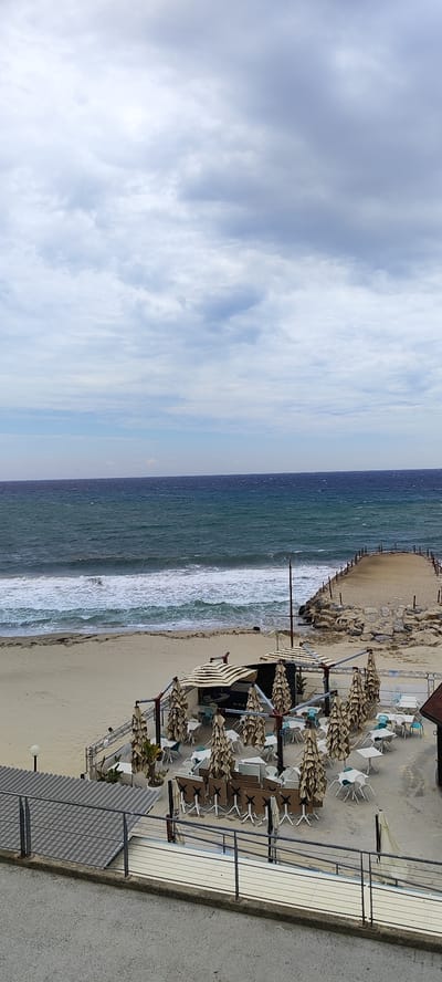 The image depicts an overcast day at a coastal area, likely a beachside restaurant in Sanremo, Italy. In the foreground, there is a patio area with numerous tables and chairs, covered by closed striped umbrellas. The patio is surrounded by a railing and a building with a grey tiled roof. Beyond the patio, there is a sandy beach with waves breaking along the shoreline. Further in the background, the sea extends to the horizon under a cloudy sky. A rocky breakwater juts out into the water on the right side of the beach. There are no people visible in the image. The scene appears calm, but the presence of the umbrellas and rough seas suggest potential inclement weather.