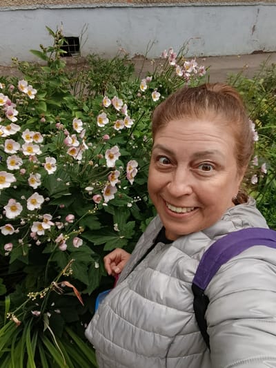 A woman with reddish-brown hair pulled into a ponytail takes a selfie outdoors, with a wall and plants behind her. She is smiling broadly, her eyes wide open and looking at the camera. She is wearing a light grey puffer jacket with a purple strap from a backpack visible over her shoulder. The background consists of a rough-textured light blue and grey wall with a small, barred window. In front of the wall is a dense cluster of green foliage and delicate, pale pink flowers with yellow centers. Some of the flowers are fully open, while others are still buds. The scene suggests an outdoor setting, possibly a garden or the side of a building. The location is stated to be Voskresensky District, Russia.
