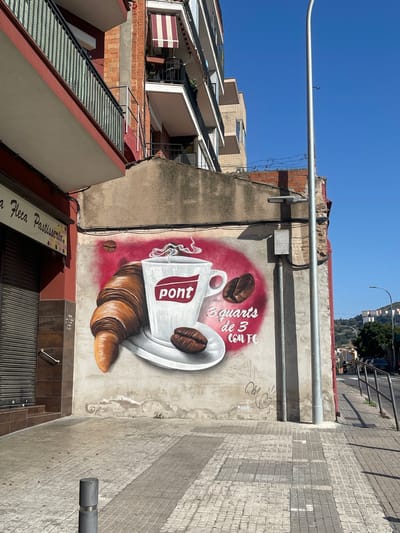 The image displays a mural painted on the side of a building in Santa Coloma de Gramenet, Spain. The mural features a large croissant, a cup of coffee with steam rising from it, and two coffee beans, all rendered in a realistic style. The coffee cup has the word "pont" in red letters inside a red rectangle. Below the coffee cup, text in white reads "3 quarts de 3 con fe". To the left of the mural is a closed storefront with a rolling metal shutter and a sign above it that reads "a fleca Pastisseria". The building facade is a mix of brick and plaster. In the background, a street with parked cars and a pedestrian crossing is visible. A tall metal lamppost stands in front of the mural. The sky is clear and blue.