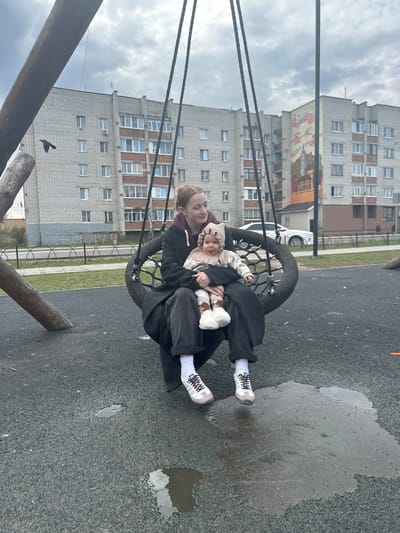 A woman holding a baby sits in a tire swing in Pokrov, Russia. A multi-story brick building looms in the background. The playground surface is wet, with puddles reflecting the sky. The woman wears a dark coat and light sneakers with white socks. The baby wears a bonnet and is bundled in light-colored clothing, holding a stuffed animal. A bird flies near the swing's wooden supports.