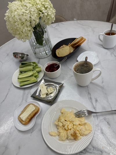 A table set for breakfast is shown in an indoor setting, likely a dining room or kitchen. The table has a marble-like surface. On the table are several food items and drinks. A white mug contains coffee with a spoon, and another white mug holds tea with a spoon. There's a small white dish with slices of toasted bread, a black oval dish with more bread slices, a white oval plate with sliced cucumbers, and a small white bowl filled with red jam. A small silver tray holds several pieces of butter. Another white dish has a slice of toast with butter. A white plate on the right contains scrambled eggs, with a fork resting on it. In the background, a glass vase holds a large bouquet of white hydrangeas. The scene is brightly lit, suggesting natural or artificial light sources. There are no people visible in the image. The specific location in Nizhnyayarkeyevo, Russia, cannot be determined from the visual information provided.