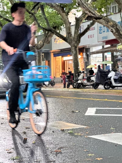 In the city of Changning District, China, on a wet street, a person rides a bright blue bicycle with a blue basket. The person, wearing a dark shirt and dark pants, is partially obscured by motion blur. The bicycle's front wheel is a lighter wood color, and the back wheel is dark. The street is wet and reflective, with scattered leaves and what appear to be raindrops. In the background, several parked electric scooters are visible. Some people in the background are holding umbrellas, one yellow and one green, indicating it might be raining. Buildings with storefronts line the street. A street sign with Chinese characters is visible. The scene suggests an urban environment during or after a rain shower.