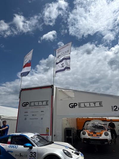 The image shows two racing cars near a GP Elite tent. One car, number 33, displays the names E. Pisani and SCAM, while the other has a number 26 on it. Flags of Israel and France are on display. The tent prominently features the GP Elite logo and the phrase "Drive to Perfection," along with sponsor logos. A partially visible mechanic works on one of the cars. The sky is blue with scattered clouds.