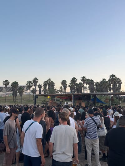 The image depicts a large outdoor gathering, likely an event or festival, taking place on a sandy area in Sant Adrià de Besòs, Spain, specifically identifiable by the large, multi-tiered concrete building resembling structures found at Parc del Fòrum. The time of day is late afternoon or early evening, indicated by the clear, light blue sky transitioning to a pale orange hue near the horizon, with a faint crescent moon visible centrally in the upper sky. In the background, a prominent, modern, grey concrete building with several levels and angular architecture is visible on the left. A retaining wall in front of the building displays graffiti. Numerous tall palm trees are distributed across the mid-ground, extending from behind the building to the right side of the frame, separating the background structures from the crowd. A central temporary structure serves as an event focal point, possibly a bar or DJ booth. It features a light-colored, possibly thatched or wooden, flat roof supported by poles, adorned with string lights and what appears to be a reflective disco ball element on its left side. A partially visible sign reading "UBOISE" is located on the structure's front. To the right of this structure, a dark blue tarp or canopy is stretched, and various wooden, basket-like decorative items hang from the roof. The foreground and mid-ground are densely populated by several hundred individuals standing on sandy ground. People are dressed in casual attire, including t-shirts, shorts, dresses, and various hats (baseball caps, bucket hats). Many individuals are holding clear plastic cups or glasses containing beverages. In the immediate foreground, from left to right: 1. A male with short dark hair, wearing sunglasses, a dark baseball cap, a grey-brown t-shirt, and brown cargo-style pants. He carries a dark cross-body bag over his left shoulder. 2. A male, seen from the back-right, with short light brown hair, wearing a white t-shirt and dark shorts. A blue cross-body strap is visible over his left shoulder. He has a small, dark triangular tattoo on his right arm and holds a clear glass with an amber liquid (likely beer) in his left hand. 3. A male, seen from behind, wearing a black and white horizontal striped t-shirt and light-colored long pants. He carries a white tote bag over his left shoulder, which has the word "Piña" and additional text printed in blue. Other crowd members show varied engagement, with some