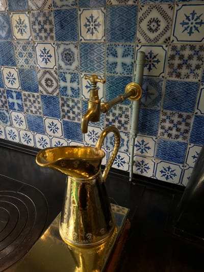 This image depicts a section of the kitchen in Claude Monet's house, located in Giverny, France. The scene is dominated by a wall covered in an array of blue and white patterned ceramic tiles. These tiles feature various intricate designs, including floral, geometric, and star-like motifs.

A brass faucet, equipped with a cross-shaped handle, extends from the tiled wall. Directly beneath the faucet, a polished, golden-hued brass pitcher with a textured, ornate handle stands on a reflective golden platform. The pitcher exhibits etched or embossed patterns around its body. This golden platform is situated on a black, solid surface, identifiable as a stove top or cooking range due to visible circular depressions that suggest burner plates. To the right of the faucet, a grey metal pipe is mounted vertically against the tiled wall, secured by two clamps. No people are present in the scene.