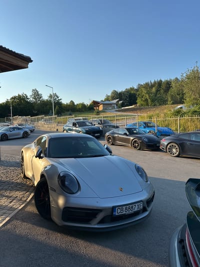 The image shows a gathering of several Porsche cars parked in an outdoor lot. The cars are of various colors including silver, dark gray, and blue. In the foreground, a silver Porsche 911 is prominently displayed, with its front end angled towards the viewer. The car has a license plate that reads "CB 8887 XE". Behind it, other Porsches are parked in a line. The setting appears to be a road or parking area adjacent to a building with a wooden roof structure on the left. In the background, there are trees and a clear blue sky, suggesting it is daytime and fair weather. The location is identified as the city of Kerenite, Bulgaria.