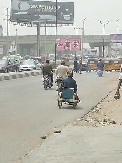 This image depicts a bustling street scene in Daurawa, Nigeria, under a hazy daylight sky. The foreground features a paved road with a distinct median barrier separating traffic lanes.
In the immediate foreground, a man drives a motorcycle from the viewer's perspective, pulling a two-wheeled, light blue and yellow passenger cart. He wears a light-colored cap and shirt. To his left, another man on a motorcycle, dressed in a dark olive-green shirt, proceeds in the same direction. Further right and slightly behind, a third man on a motorcycle, in a dark shirt, is visible. On the unpaved shoulder in the lower right, a human arm and hand are visible, holding a dark, spherical object. Two dark, irregular objects lie on the unpaved shoulder near the bottom edge.
The mid-ground shows a mix of vehicles. A dark-colored sedan and a silver sedan are visible in the left lane. On the right side of the road, several yellow motorized tricycles, commonly known as Keke Napeps, are either parked or moving slowly. Several pedestrians are present on or near the road, including one individual in a dark shirt walking left-to-right across the street.
The background is dominated by an elevated concrete bridge or overpass. Above the bridge, the sky appears overcast or hazy. Two prominent billboards are visible: a large black "SWEETHOME BATHROOM" advertisement depicting bathroom fixtures on the left, and a red "SAY YES TO THE TASTE YOU LOVE!" billboard featuring a red beverage bottle in the center. Multiple tall streetlights with apparent solar panels are positioned along the road. Traffic continues into the distance.
