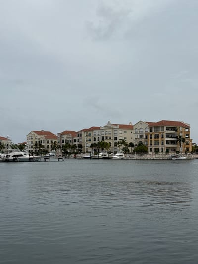 This media file depicts a waterfront scene in Higüey, Dominican Republic, likely a resort or residential area. The primary subjects are several multi-story buildings with red-tiled roofs and cream-colored exteriors, featuring arched windows and balconies. These buildings appear to be condominiums or apartments, arranged in a row along a marina.
In the foreground, several white yachts and boats are docked along a pier. The water in the marina is calm, with visible ripples and reflections. Palm trees are scattered between the buildings and along the waterfront, adding a tropical element to the scene. The sky is overcast with light gray clouds, suggesting a cloudy or potentially rainy day. The overall mood is serene and tranquil, typical of a coastal setting.
There is no visible text in the image. The time of day is indeterminate due to the overcast sky, but it appears to be daytime. The location cues, such as the architecture, palm trees, and boats, combined with the provided location context, strongly suggest a Caribbean setting. No people are clearly visible in the image, and no specific activity or event is taking place beyond the boats being docked. The focus is on the architecture and the waterfront landscape.