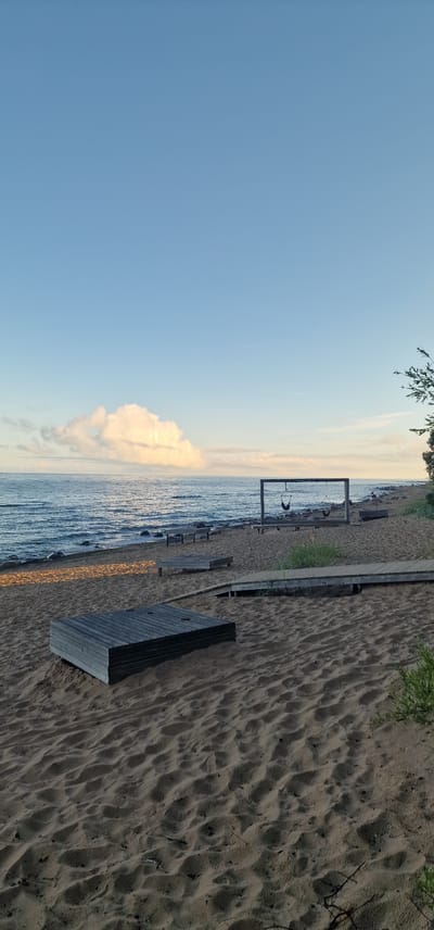 A calm, wide shot of a beach in Vārzas, Latvia, shows a calm sea with small waves, and a sandy beach in the foreground. The sky is a clear blue with a large, fluffy white cloud on the horizon. In the middle ground, a wooden swing set with two swings is visible, positioned facing the sea. Several wooden benches are scattered on the sand near the water's edge, and a wooden walkway leads towards the swing set. In the foreground, a low, square wooden platform rests on the sand. The sand is textured with ripples and shadows, suggesting it is late afternoon or early morning. A single tree branch is visible on the right edge of the frame, adding a touch of nature to the scene. The overall impression is one of tranquility and natural beauty.