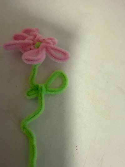 A pink and green pipe cleaner flower is positioned diagonally on a textured, light-colored surface, with the flower's bloom at the top left and its stem extending towards the bottom right. The flower is constructed from fuzzy pipe cleaners, with pink forming the petals and a small green piece at the center. The stem is green and has a curved loop. The background is a plain, slightly mottled surface, possibly a table or wall. The image has a grainy quality and is lit from above. The location is specified as Ridabu, Norway.
