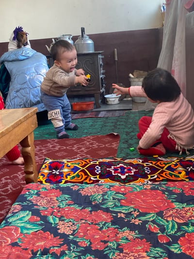 A toddler lying on a blue quilted blanket holds a yellow toy, while another child in a pink sweater reaches for something near a metal stove. The room features a vibrant floral print blanket or fabric. A section of intricate patterned rug is visible. The scene is cozy, likely indoors, with simple furnishings.