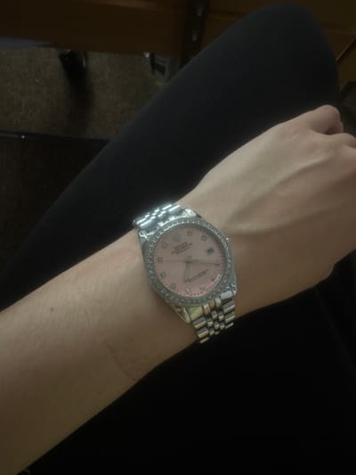 A close-up, eye-level shot shows a person's left forearm, wearing a silver Rolex Datejust watch with a pink dial and diamond bezel. The watch is positioned on the wrist, with the bracelet clearly visible. The person is wearing black clothing that covers their arm and leg. The background is blurred and dark, with hints of wooden furniture and a carpeted floor. No location in Yessentuki, Russia, is discernible from the image.