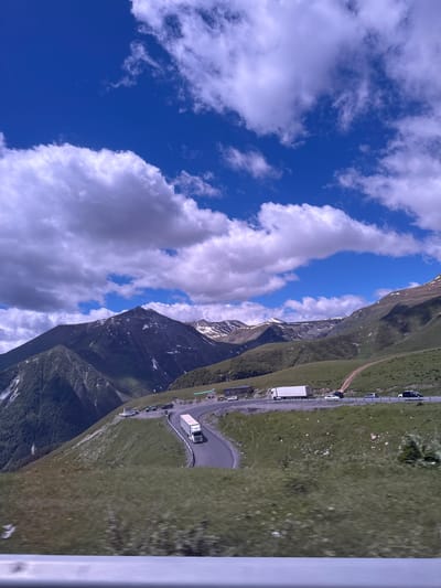 The image captures a sweeping view of a mountainous landscape in Gudauri, Georgia, under a bright blue sky dotted with fluffy white clouds. A winding asphalt road curves through the scene, with a white semi-trailer truck driving on it. Several cars are parked or driving along the road, and a small, dark-roofed building is visible on the left side. The slopes of the mountains are covered in lush green grass, with patches of snow appearing on the higher peaks. The foreground is blurred, suggesting the photo was taken from a moving vehicle. The overall impression is one of a scenic mountain pass on a clear day.