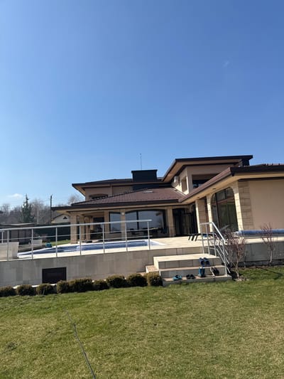 The image displays the exterior of a multi-level residential property in Erden, Bulgaria, under a clear blue sky. The building features light beige walls, a dark brown tiled roof, and extensive glass windows and doors. A black, rectangular structure topped with an antenna is present on the roofline. An outdoor swimming pool with blue mosaic tiling occupies a raised, light-colored tiled patio area, partially enclosed by a stainless steel railing. Steps, appearing to be wood integrated into a stone wall, descend from the patio to a meticulously maintained green lawn. Four pairs of footwear—two pairs of flip-flops, one pair of blue clogs, and one pair of dark shoes—are neatly arranged on the lower steps. A row of low, trimmed evergreen shrubs borders the patio's base. A dark cable or hose lies across the foreground lawn. In the background, dormant deciduous trees and glimpses of other residential structures, alongside utility poles and lines, are visible. No individuals are present in the scene.