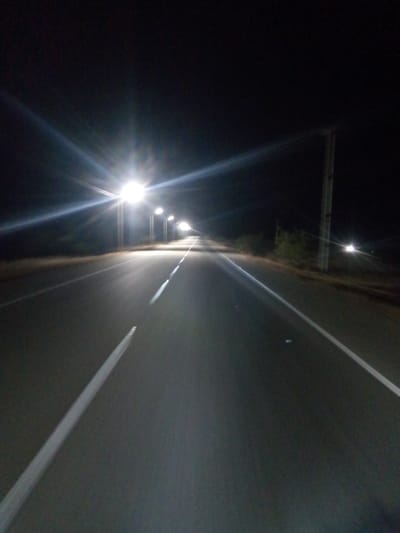 The image depicts a paved road at night, lit by a series of streetlights extending into the distance. White lines mark the lanes. The road is likely located in or near El Dátil, Venezuela. There are sparse trees/foliage visible on either side of the road. In the distance, the last street light can be seen.